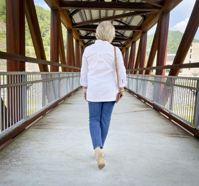 Over 40 Fashion Blogger, Tania Stephens, is walking on a bridge wearing a white button down tunic shirt, ankle length jeans, and neutral kitten heels