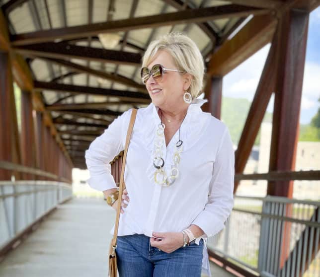 Over 40 Fashion Blogger, Tania Stephens, is wearing a white button down shirt with a large ruffle, sunglasses, a long shell necklace, and earrings