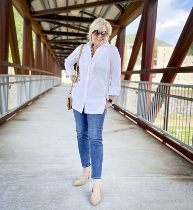 Over 40 Fashion Blogger, Tania Stephens, is in the middle of a bridge wearing a white button down shirt with a large ruffle, ankle length jeans, and neutral kitten heels