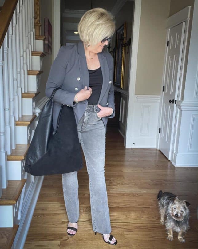 Over 40 Fashion Blogger, Tania Stephens, is styling a double-breasted gray blazer with a black bodysuit, gray raw hem jeans, black heels, and holding a large black handbag