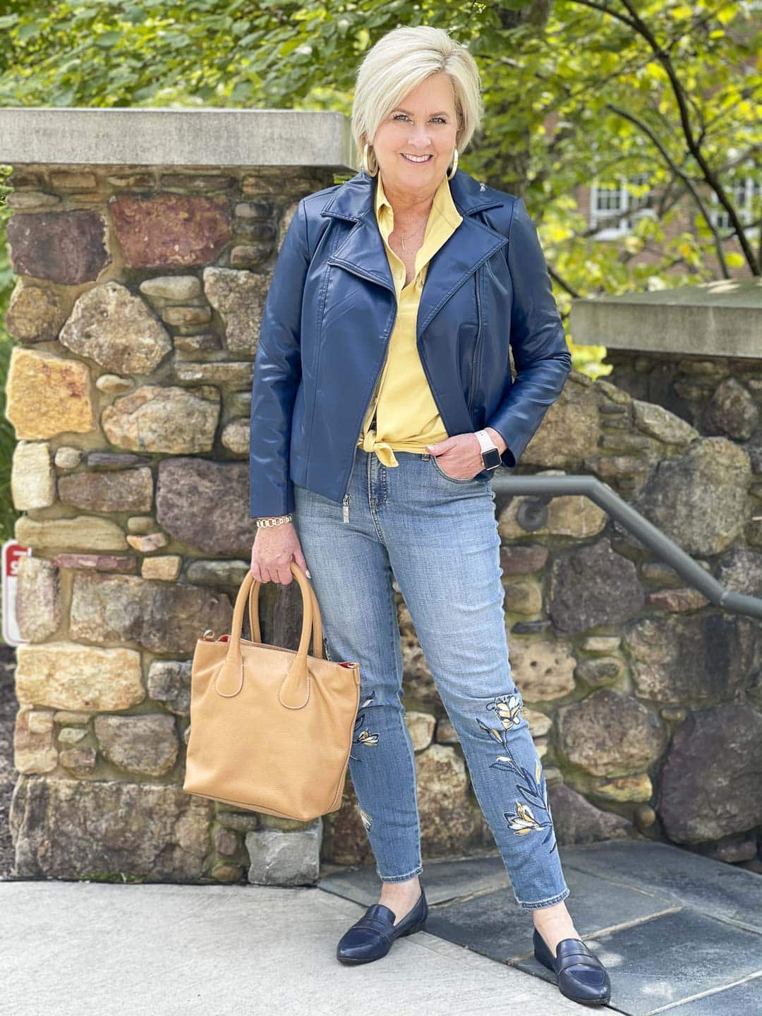 Over 40 Fashion Blogger, Tania Stephens, is wearing a navy faux leather moto jacket with a Chamois yellow button up shirt, embroidered jeans, and navy penny loafers