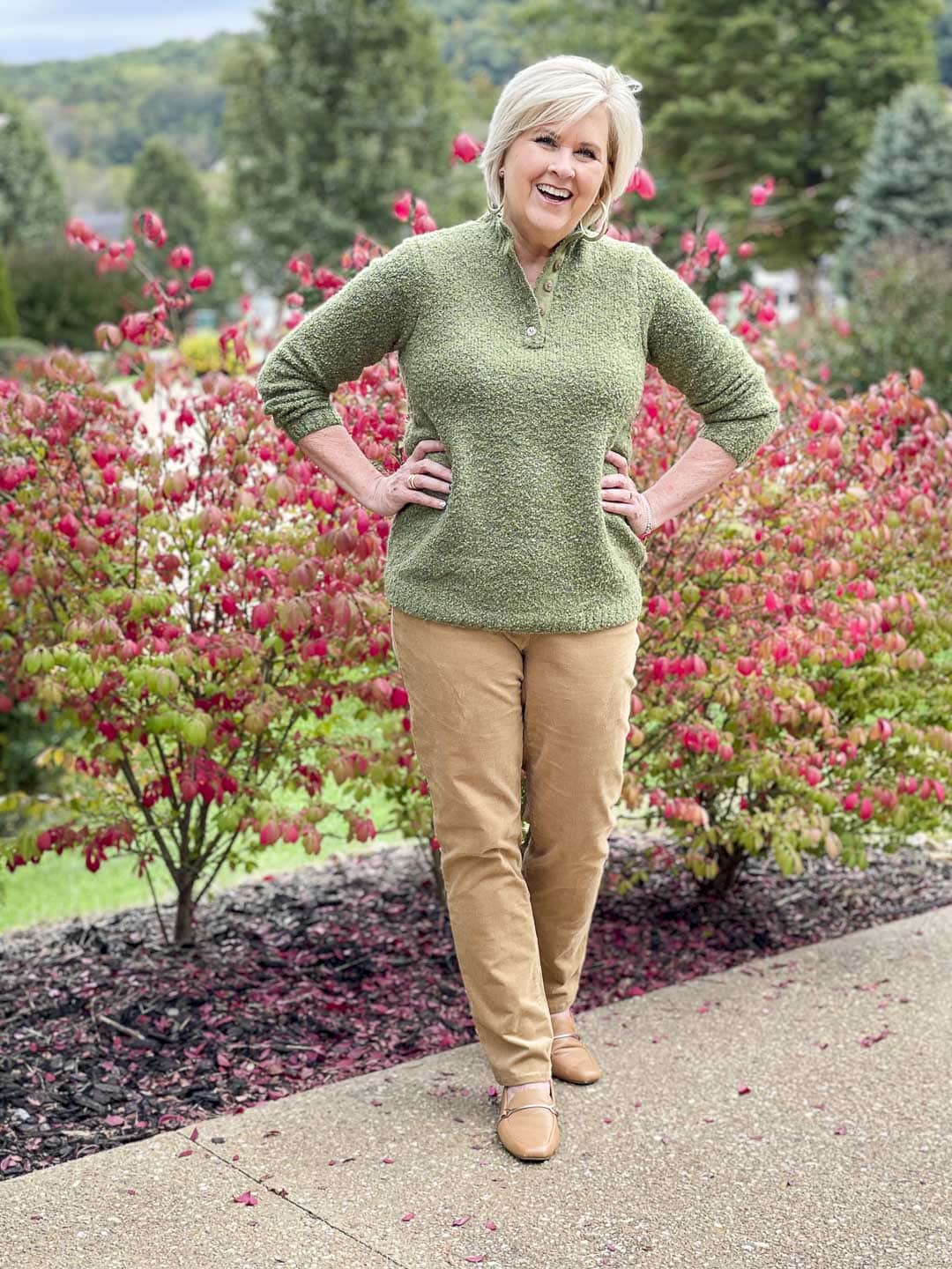 Over 40 Fashion Blogger, Tania Stephens is in front of a red bush wearing a warm and cozy fall outfit of an olive green sweater and camel corduroy pants