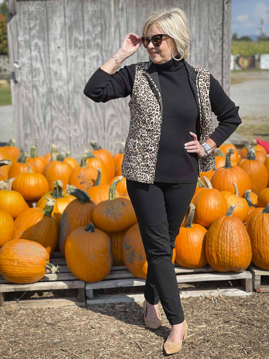 Over 40 Fashion Blogger, Tania Stephens, is in a pumpkin field wearing a reversible animal print vest, a black turtleneck, sunglasses, and black pants