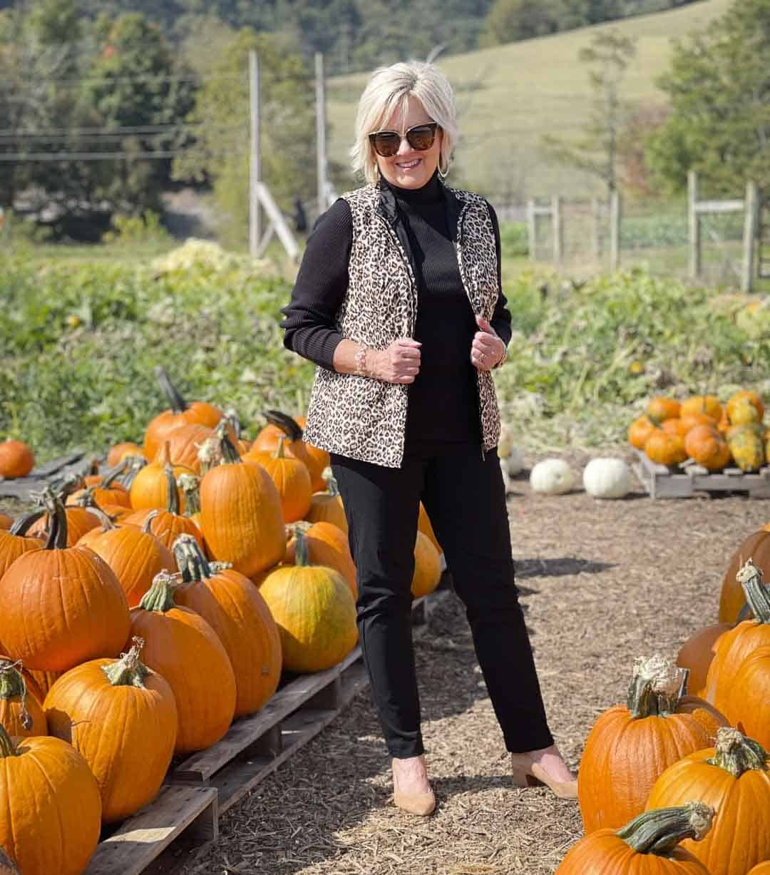 Over 40 Fashion Blogger, Tania Stephens, is in a pumpkin field wearing a reversible animal print vest with a black turtleneck and black pants