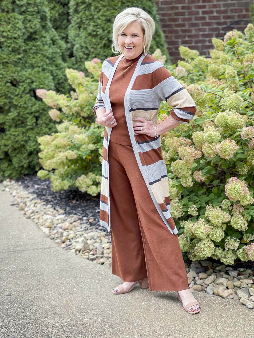 Over 40 fashion blogger, Tania Stephens, is standing by hydrangeas wearing a striped cardigan with a dark rust sweater tank and pants perfect for Thanksgiving Dinner