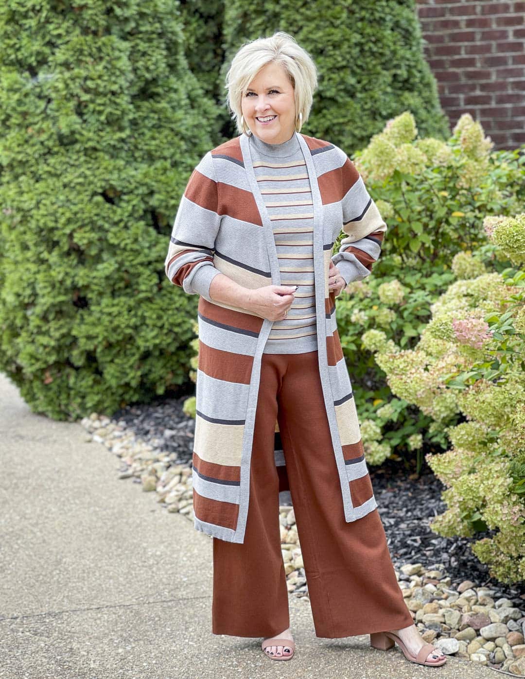 Over 40 fashion blogger, Tania Stephens, is standing by hydrangeas wearing a striped cardigan with a gray striped sweater tank and rust pants perfect for Thanksgiving Dinner