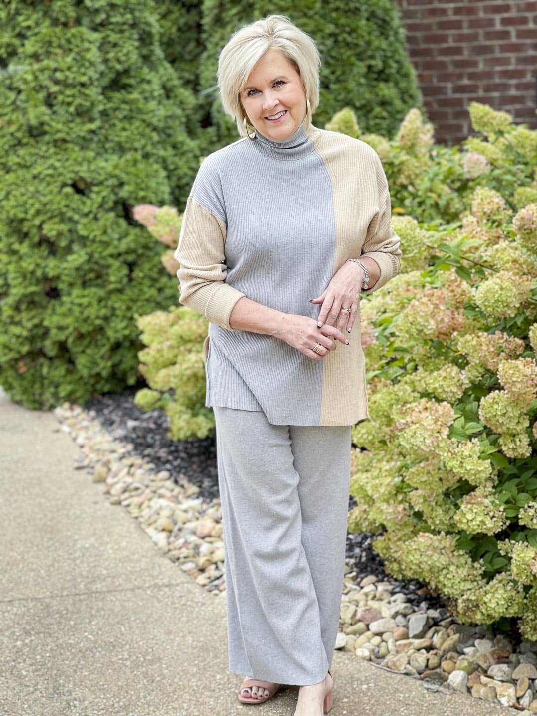 Over 40 fashion blogger, Tania Stephens, is standing by hydrangeas wearing a gray color block sweater and gray pants perfect for Thanksgiving Dinner