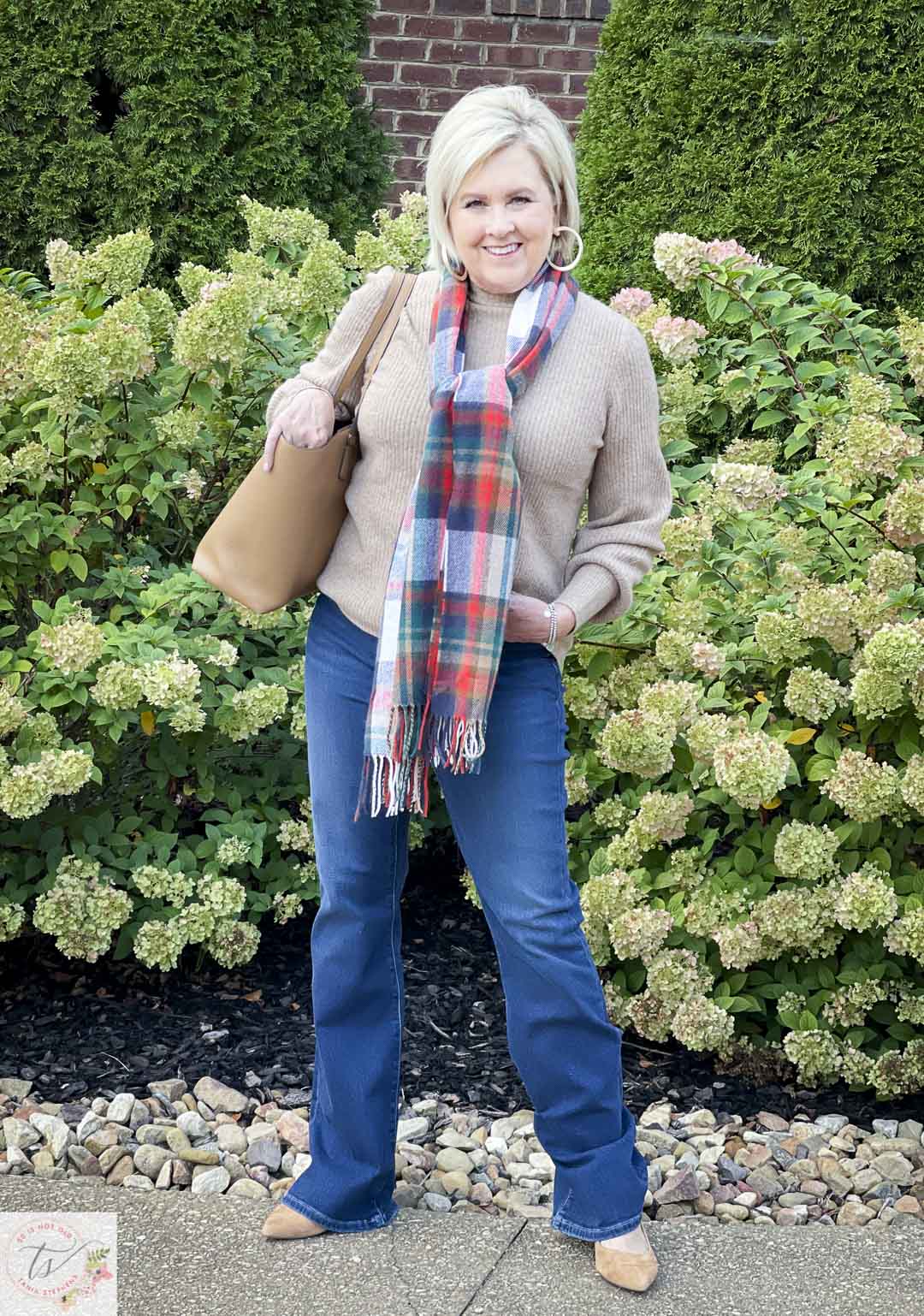 Over 40 Fashion Blogger, Tania Stephens, is wearing a ribbed mockneck sweater with a plaid scarf, mid-wash flare jeans and is carrying a Tory Burch tote
