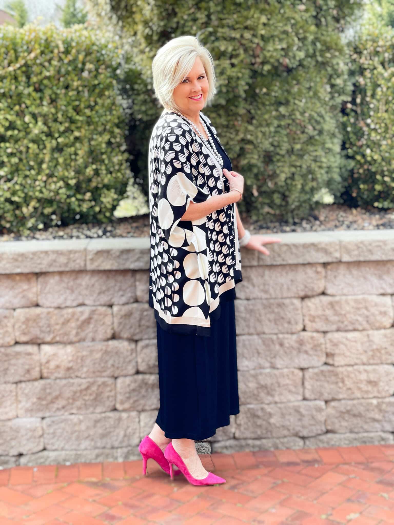 Over 40 Fashion Blogger, Tania Stephens is in front of a block wall wearing a black dress and a black and white ruana