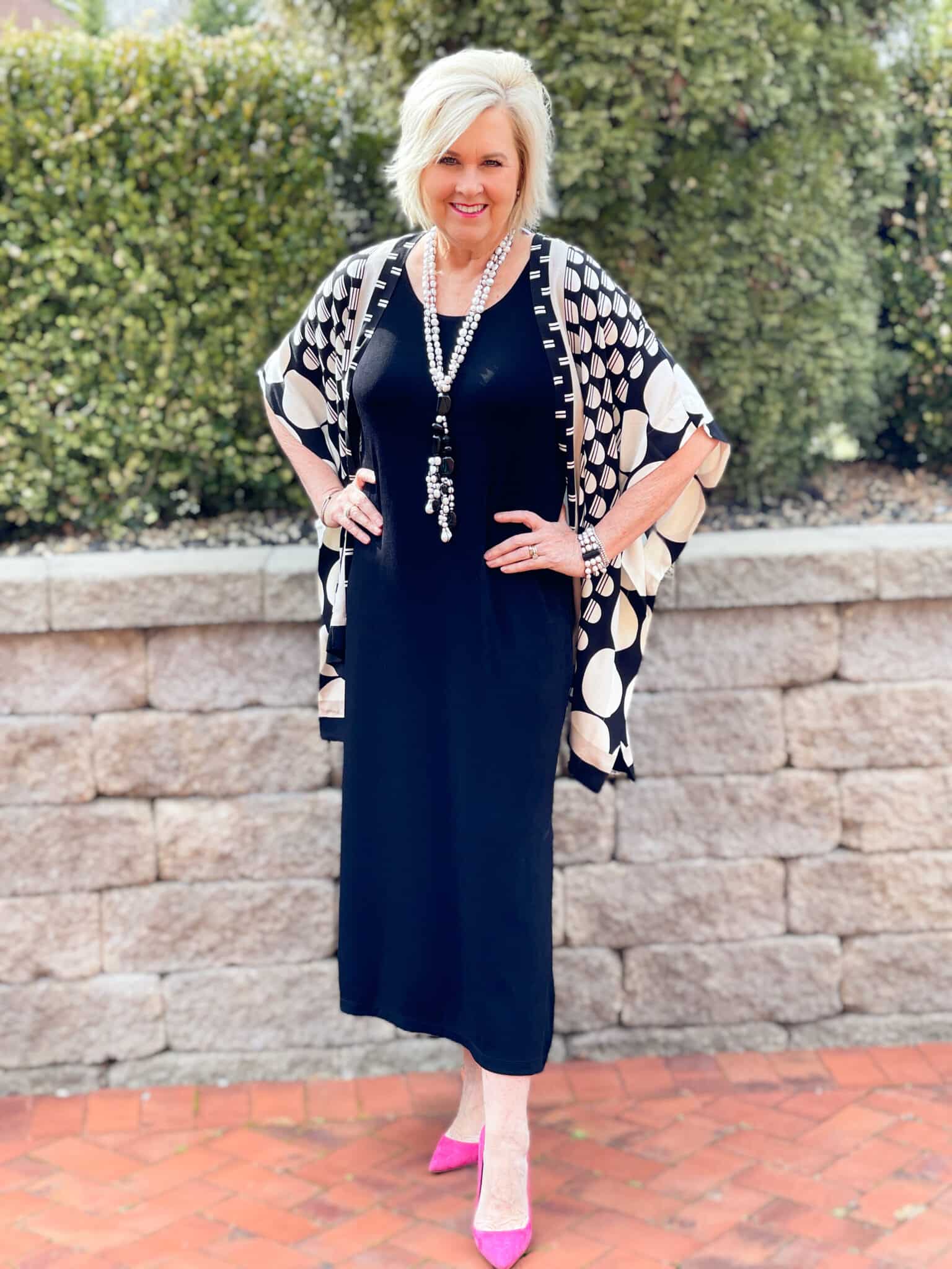Over 40 Fashion Blogger, Tania Stephens is in front of a block wall wearing a black midi dress and a black and white ruana
