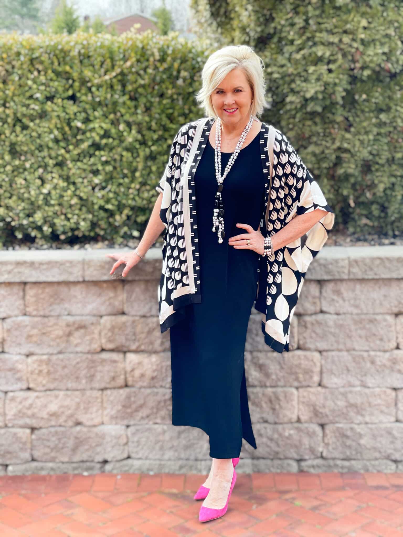 Over 40 Fashion Blogger, Tania Stephens is in front of a block wall wearing a black midi dress and a black and white ruana with pink heels