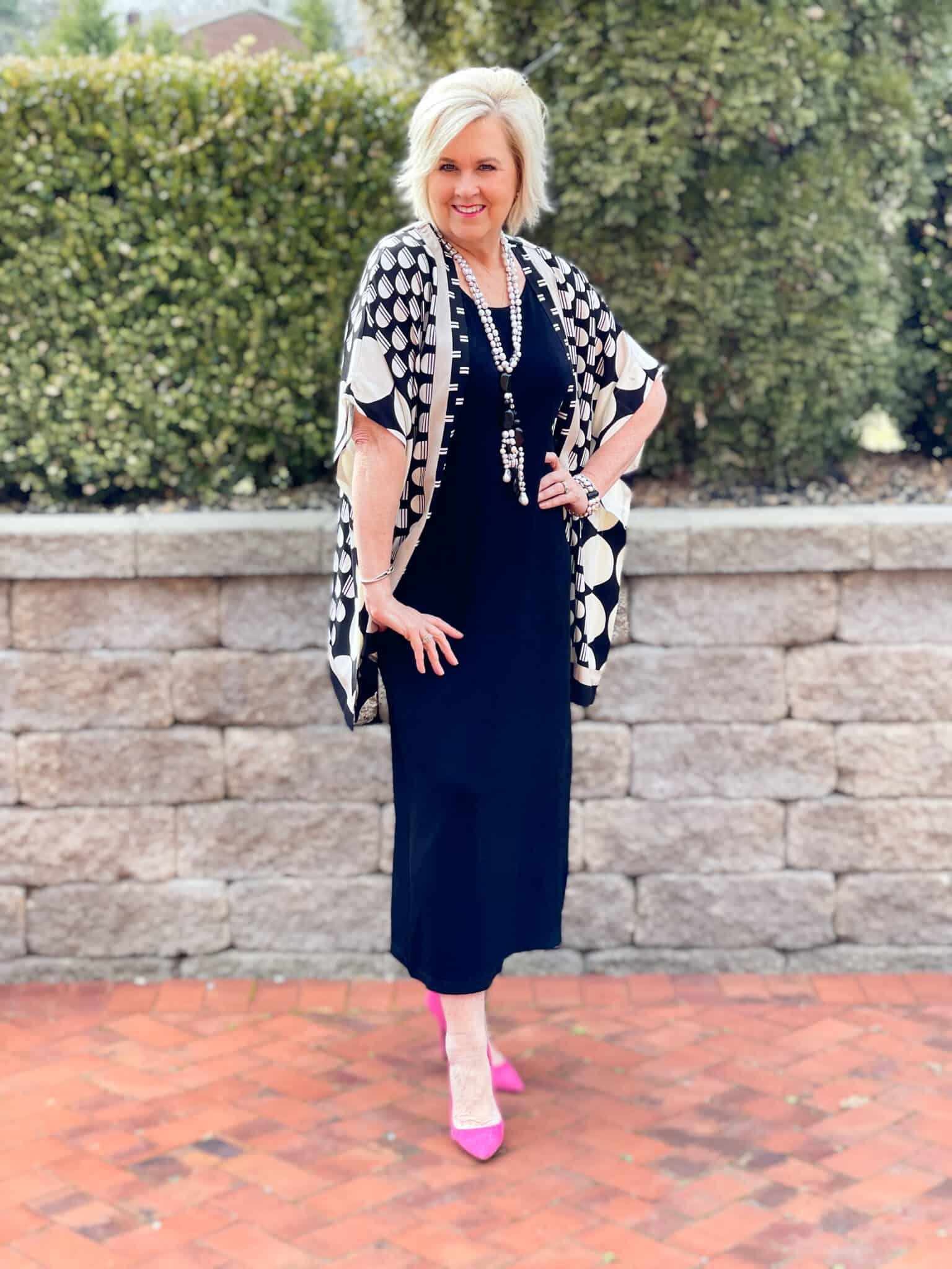 Over 40 Fashion Blogger, Tania Stephens is in front of a block wall wearing a black midi dress and a Ruana with pink heels