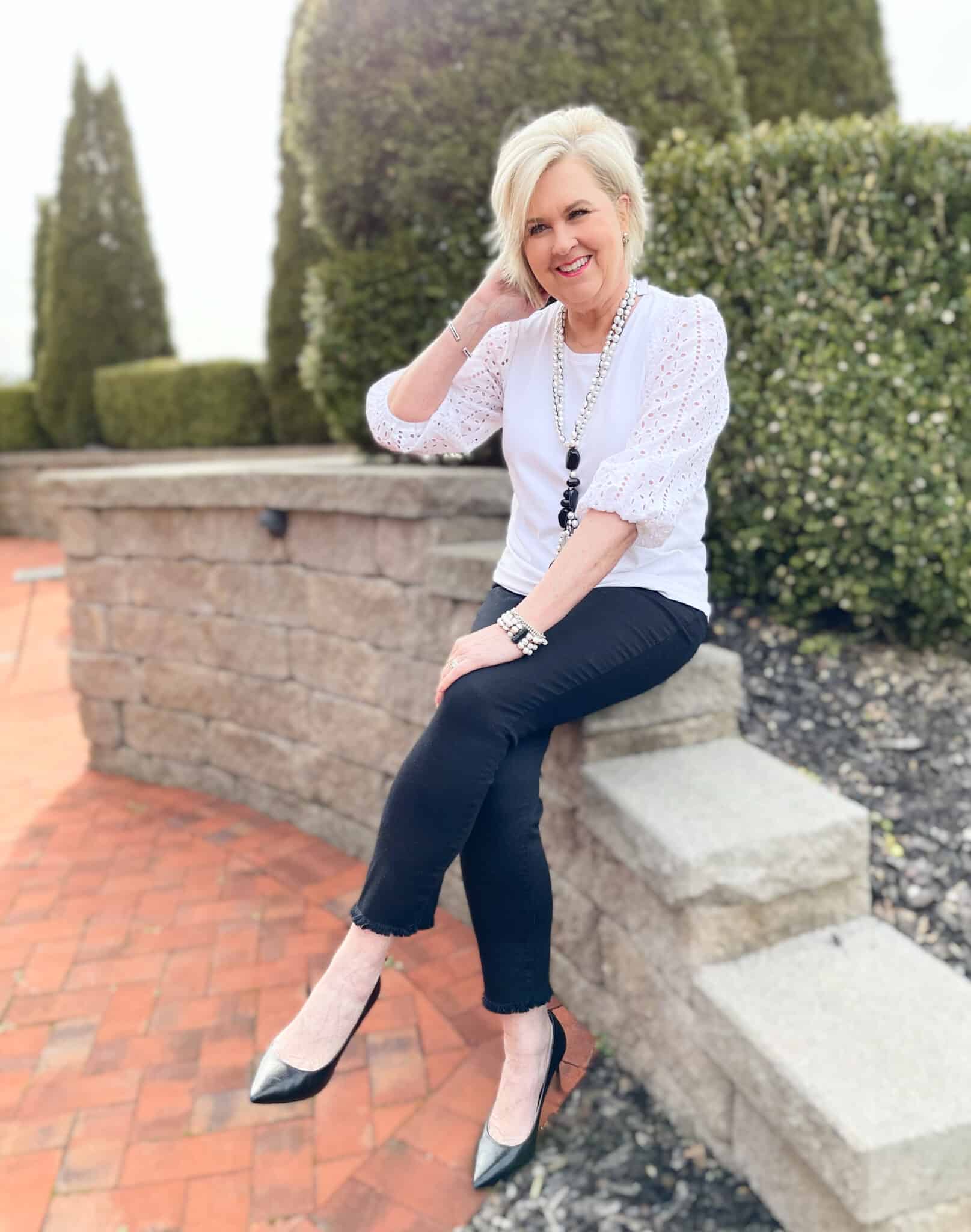 Over 40 Fashion Blogger, Tania Stephens is sitting on a block wall with her legs crossed wearing a white eyelet top and black jeans