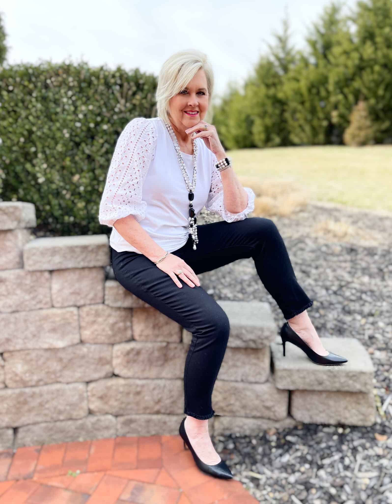 Over 40 Fashion Blogger, Tania Stephens is sitting on a block wall wearing a white eyelet top and black jeans
