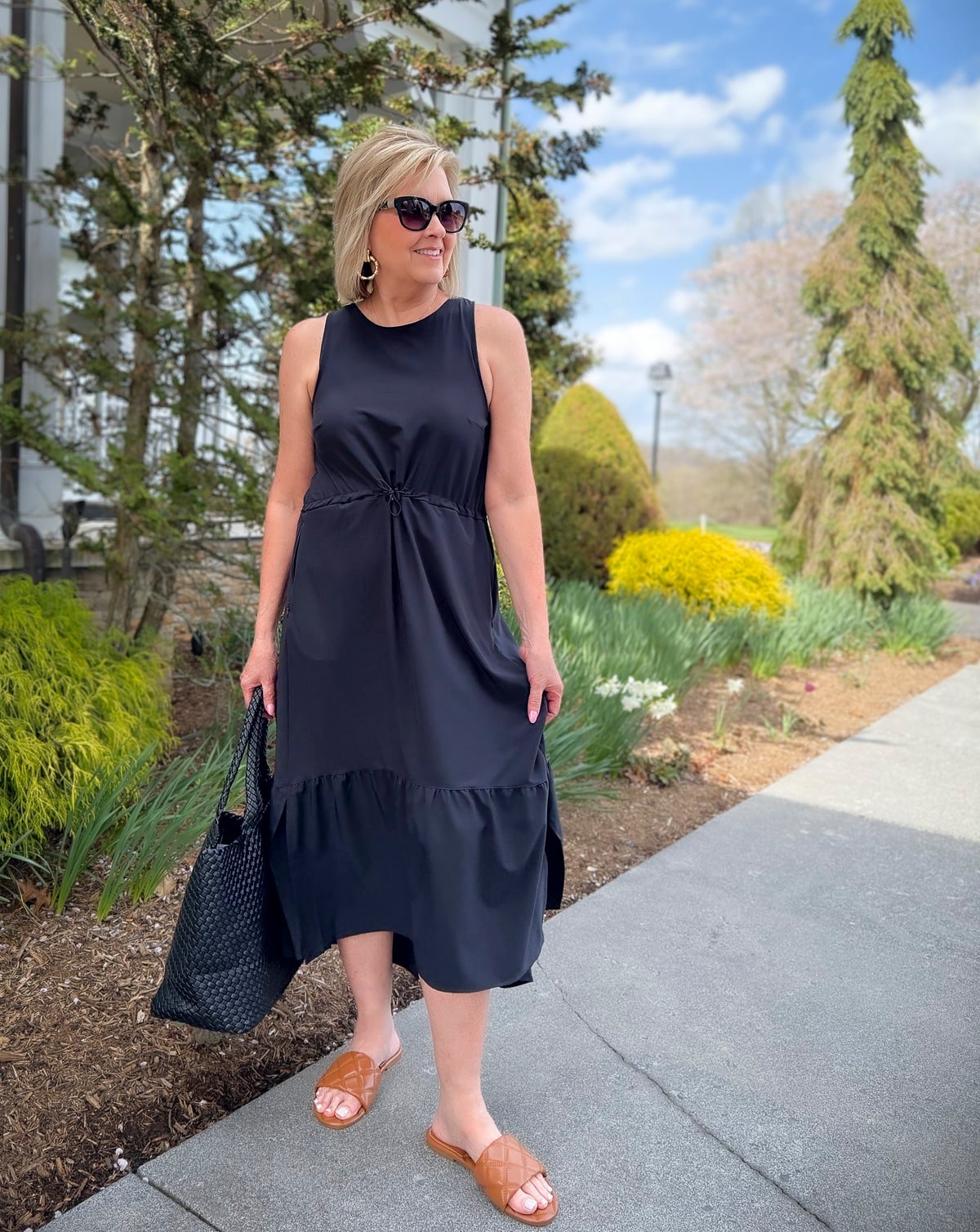 Woman wearing a sleeveless black midi dress with a woven tote and quilted slides for a relaxed spring outfit