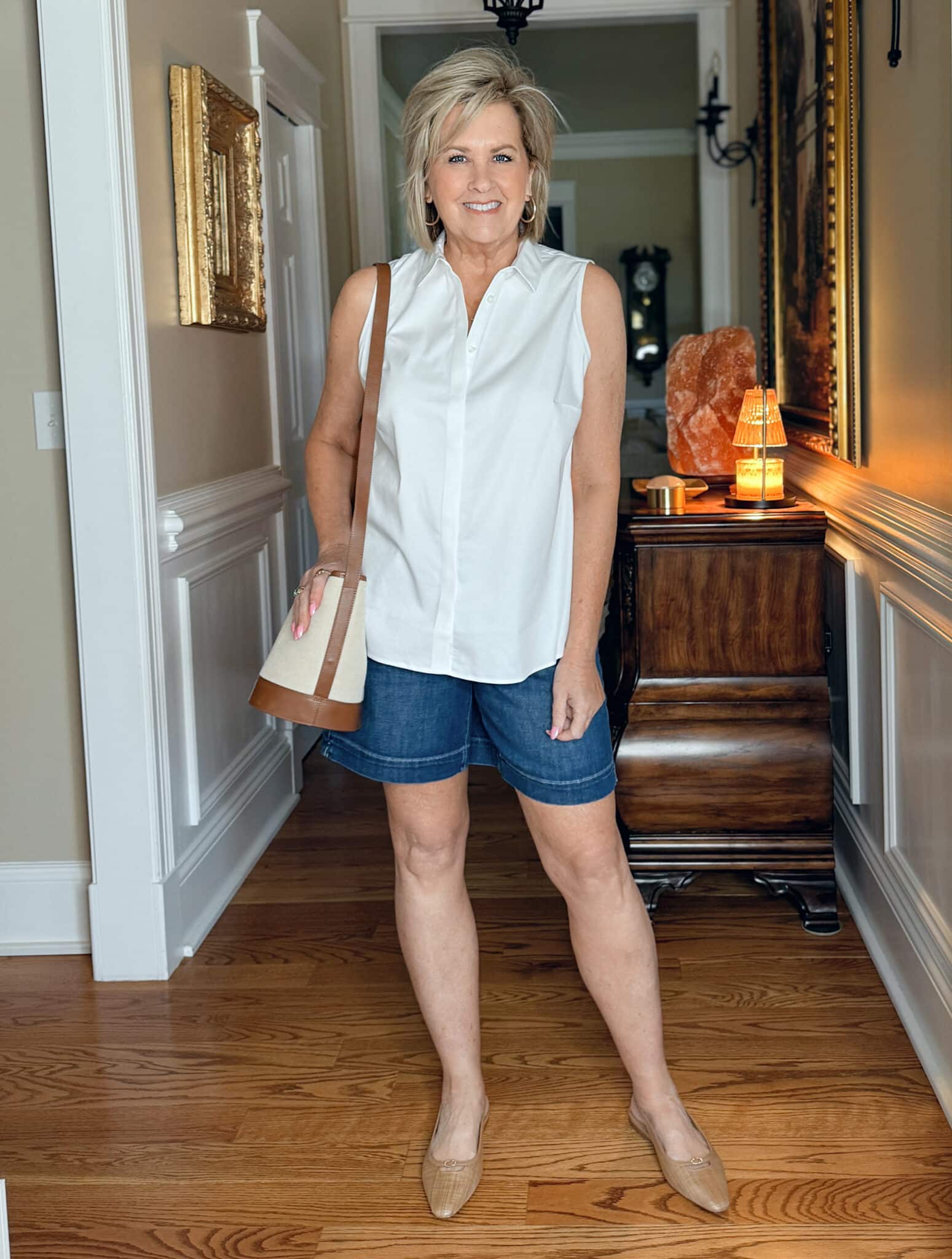 Woman over 50 wearing a sleeveless white button-up shirt with dark denim shorts, canvas crossbody bag, and flat woven sandals—styled for a casual summer weekend.