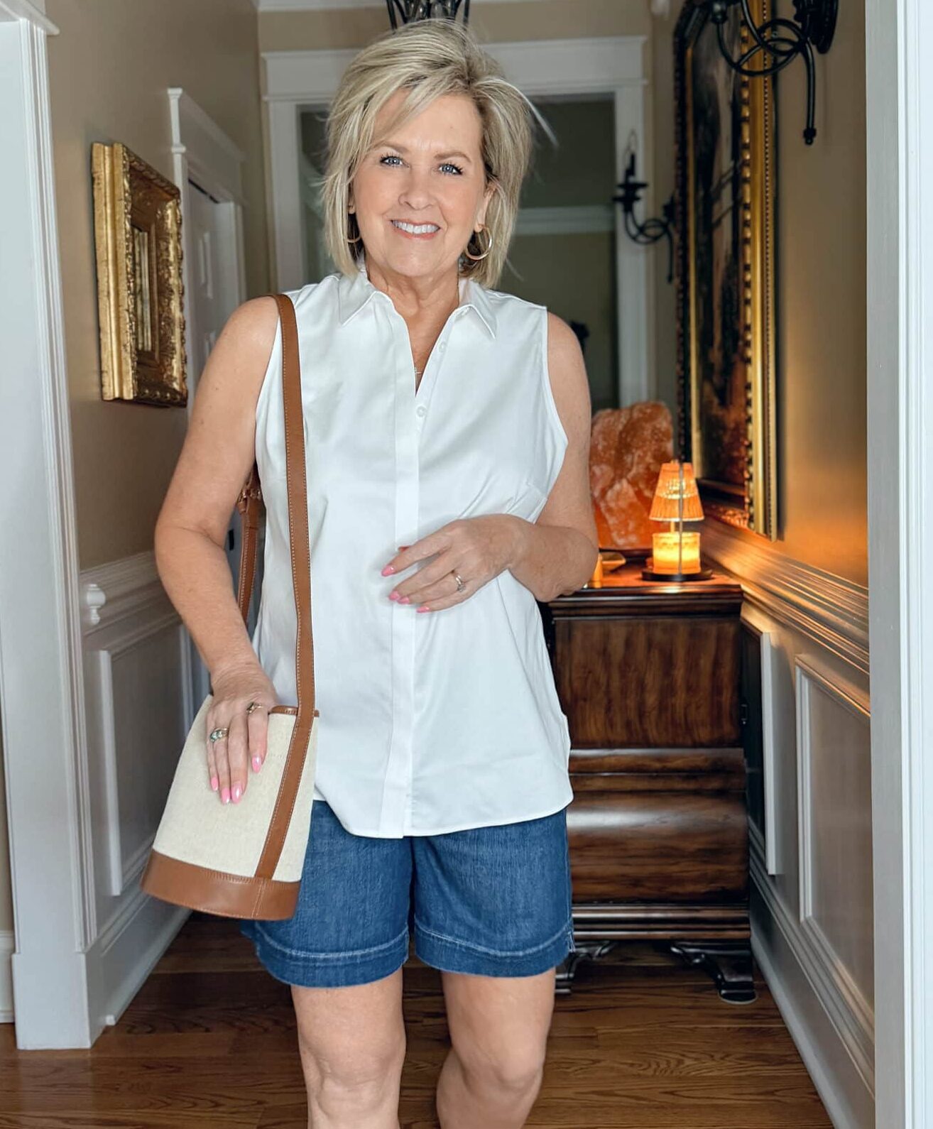 Woman over 50 wearing a sleeveless white button-up shirt with dark denim shorts and a canvas handbag