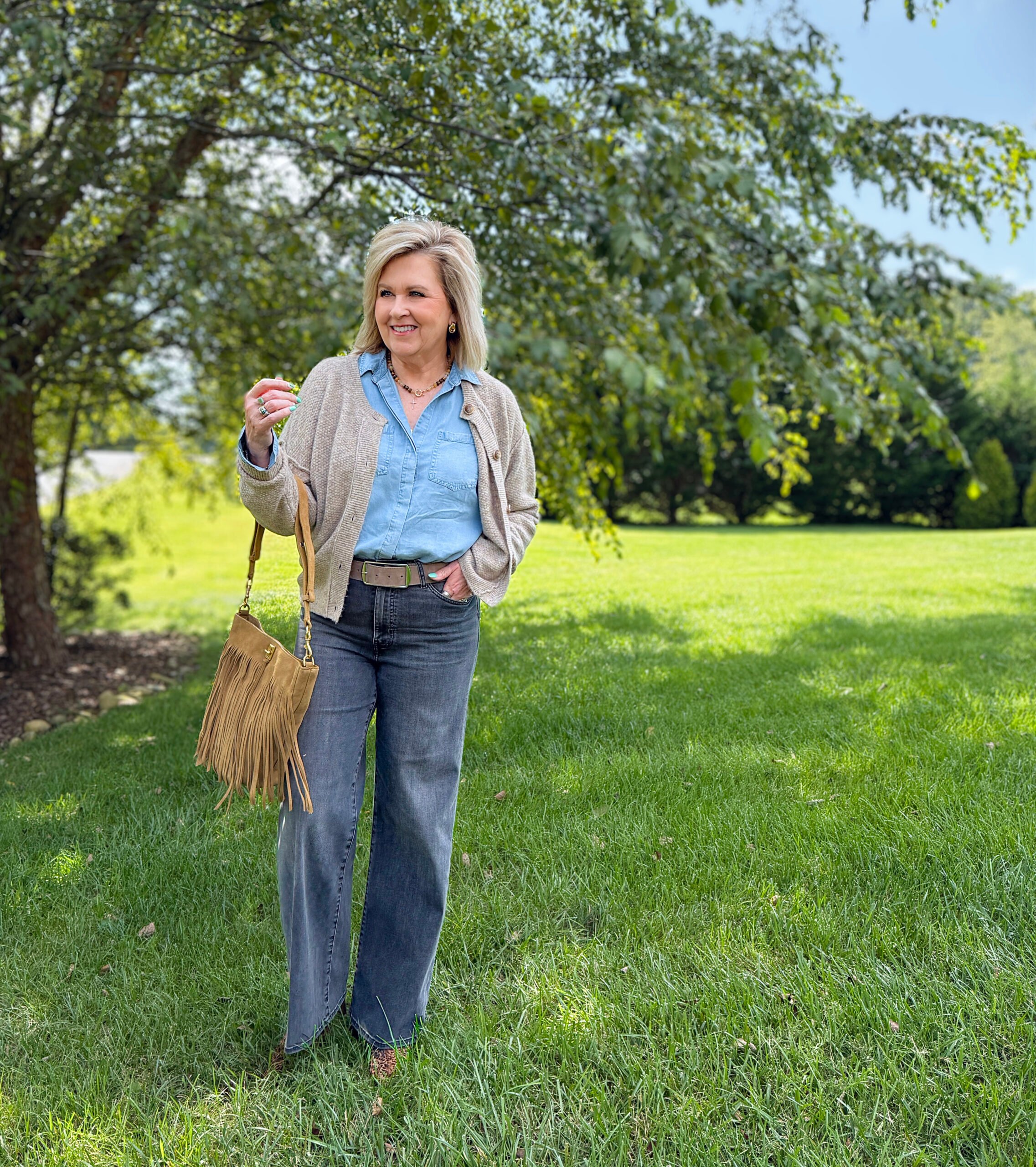 Woman over 50 wearing western-inspired fall outfit with brown cardigan, chambray shirt, black wide-leg jeans, fringe bucket bag, and calf hair booties from Nordstrom Rack.