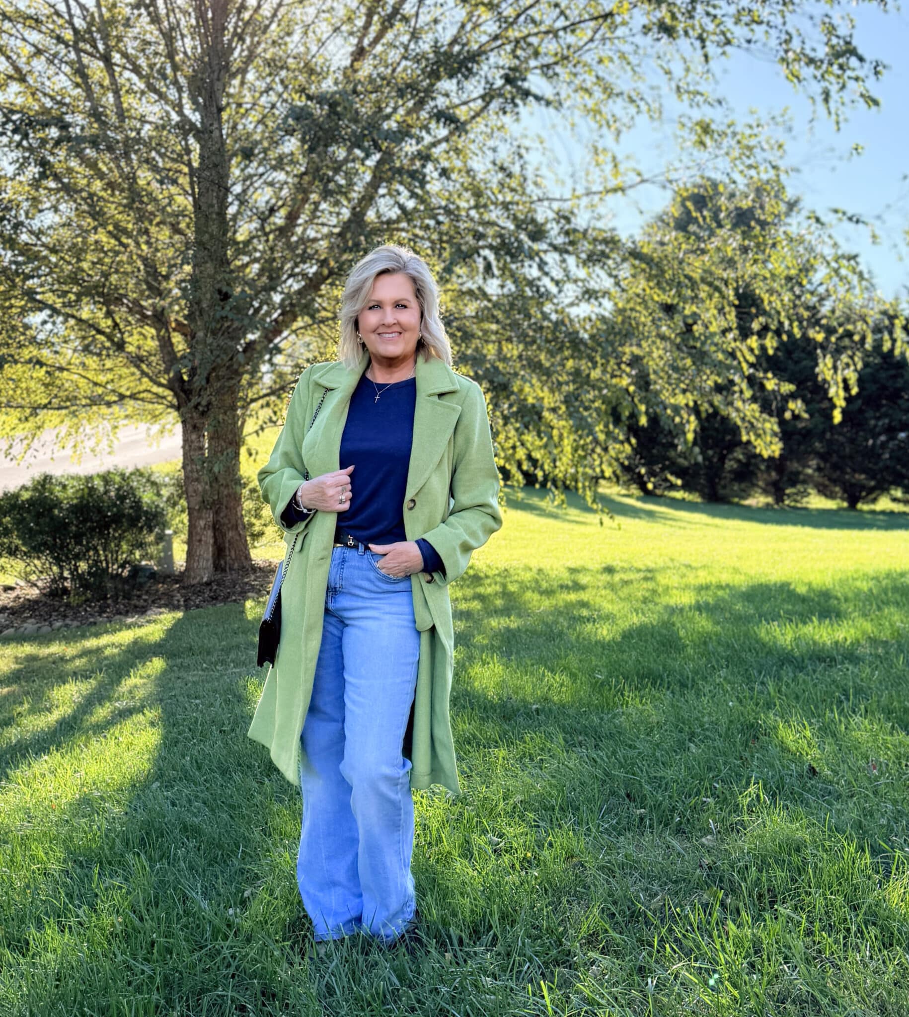 Tania Stephens standing under a tree wearing a Kohl's long green coat with a blue tee and lightwash jeans
