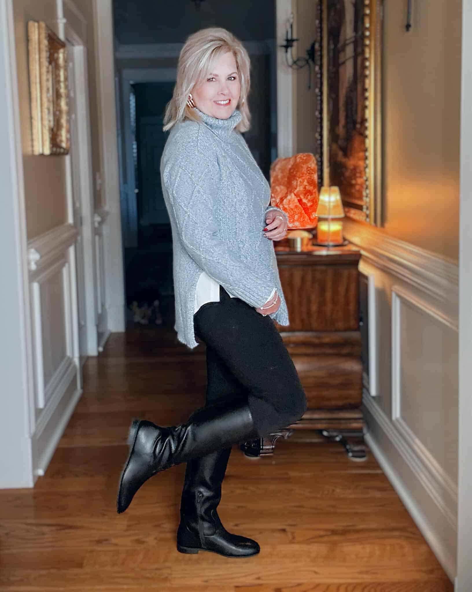 Tania showing the side view of a icy blue cable knit cowl neck sweater with black leggings and riding boots from Kohl's